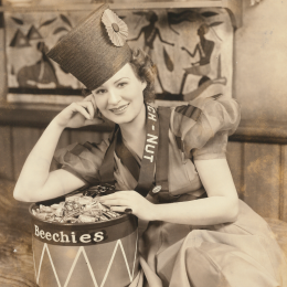 Sepia toned photo of a young woman leaning on a drum full of Beech-Nut Gum. She wears an outfit with puffed up sleeves and a talk shako hat. 