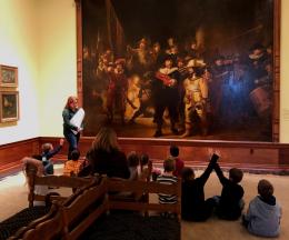 Group of students sit in front of a large painting. A facilitator stands in front of the group. 