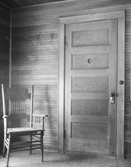 interior of a room. Walls are paneled with wood, a door has a round peep hole in it. There is a chair to the left of the door