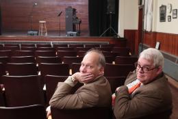 Two older men sitting in auditorium chairs looking back at the camera